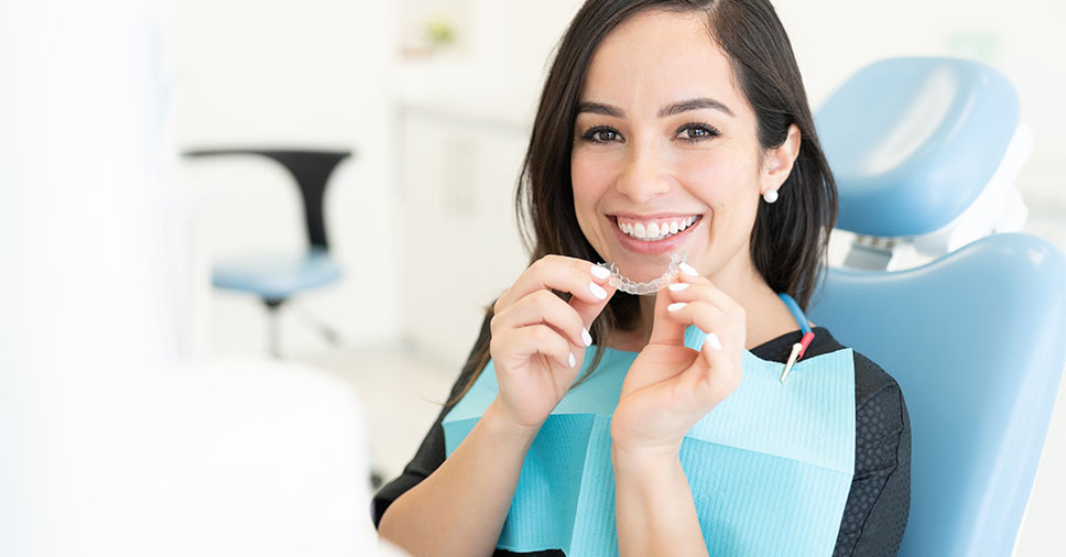 mulher sorrindo na cadeira de dentista segurando o aparelho ortodôntico transparente